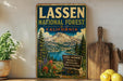 A vintage-style metal sign for the Lassen National Forest in California, featuring a scenic mountain landscape with a lake and pine trees.