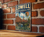 A vintage-style metal sign for Lowell National Historical Park in Massachusetts, featuring an illustration of a historic building and a river.