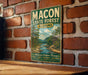 A vintage-style metal sign for the Macon State Forest in Alabama, featuring a scenic landscape with a river and forest.