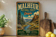 A vintage-style metal sign for Malheur National Forest in Oregon, featuring a scenic landscape with mountains, trees, and a river.
