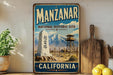 A vintage-style metal sign for the Manzanar National Historic Site in California, featuring a desert landscape with mountains in the background.