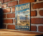 A vintage-style metal sign for the Manzanar National Historic Site in California, featuring a mountain landscape and a tower.