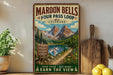 A vintage-style metal sign advertising the Maroon Bells Four Pass Loop in Colorado, featuring a scenic mountain landscape with a lake and trail.