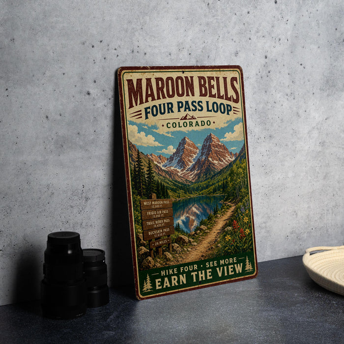 A vintage-style metal sign advertising Maroon Bells Four Pass Loop in Colorado, featuring a scenic mountain landscape with a lake and trail.