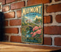 A vintage-style metal sign with the text "MAYMONT VIRGINIA" and an illustration of a bridge, flowers, and a house, mounted on a brick wall.