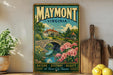 A vintage-style poster of Maymont, Virginia, featuring a scenic landscape with a bridge, flowers, and a house, displayed on a shelf next to a wooden cutting board and a potted plant.