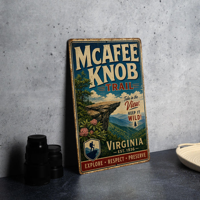 A vintage-style metal sign for the Mcafee Knob Trail, featuring a scenic mountain landscape and the text "Take in the View! Keep it Wild!" The sign is displayed on a gray concrete surface, with a stack of black camera lenses and a white dish visible in the background.