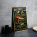 A vintage metal sign for McConnells Mill State Park in Pennsylvania, featuring a red brick building and a waterfall, is displayed on a gray countertop next to some black camera lenses.