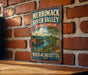 A vintage-style metal sign for Merrimack River Valley, Massachusetts, featuring a scenic landscape with a river, buildings, and trees.
