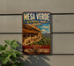 A vintage-style metal sign for the Mesa Verde National Park in the United States, featuring a scenic image of the ancient cliff dwellings in the background.