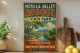A vintage-style sign for Mesilla Valley Bosque State Park, featuring a scenic landscape with mountains, trees, and a river.