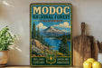 A vintage-style metal sign for Modoc National Forest, California, featuring a scenic mountain landscape with pine trees, clear lakes, and a river.