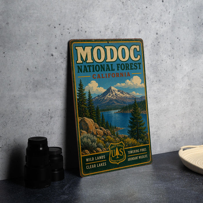 A vintage metal sign for Modoc National Forest, California, featuring a scenic landscape with mountains, forests, and a lake.