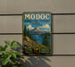 A vintage-style metal sign for Modoc National Forest, California, featuring a mountain landscape with pine trees and clear lakes.
