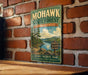 A vintage-style metal sign for the Mohawk State Forest in Connecticut, featuring a scenic landscape with a lake, mountains, and pine trees.