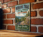 A vintage-style metal sign for the Mohawk Trail in Massachusetts, featuring a scenic mountain landscape with a winding road.