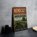 A vintage metal sign with the text "MONOCACY NATIONAL BATTLEFIELD MARYLAND" and an illustration of a cannon and a bridge, placed on a gray countertop next to some black camera lenses.
