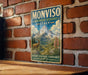A vintage-style metal sign with a mountain landscape and a goat, mounted on a brick wall.