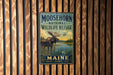 A metal sign with a moose and a lake, mounted on a wooden wall.