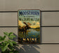 A metal sign with a moose and a river, mounted on a wall next to a plant.