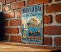 A vintage-style metal sign for Morro Bay State Marine State Recreation Area, featuring a sailboat and sea otters, is displayed on a brick wall.