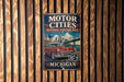 A vintage-style metal sign with the text "Motor Cities National Heritage Area" and "Michigan" displayed on it, mounted on a wooden wall.