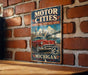 A vintage-style metal sign with the text "Motor Cities National Heritage Area" and "Michigan" displayed on it, mounted on a brick wall.