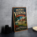 A vintage-style metal sign for Mount Vernon, Virginia, featuring a house and trees, is displayed on a gray countertop next to some black camera lenses.