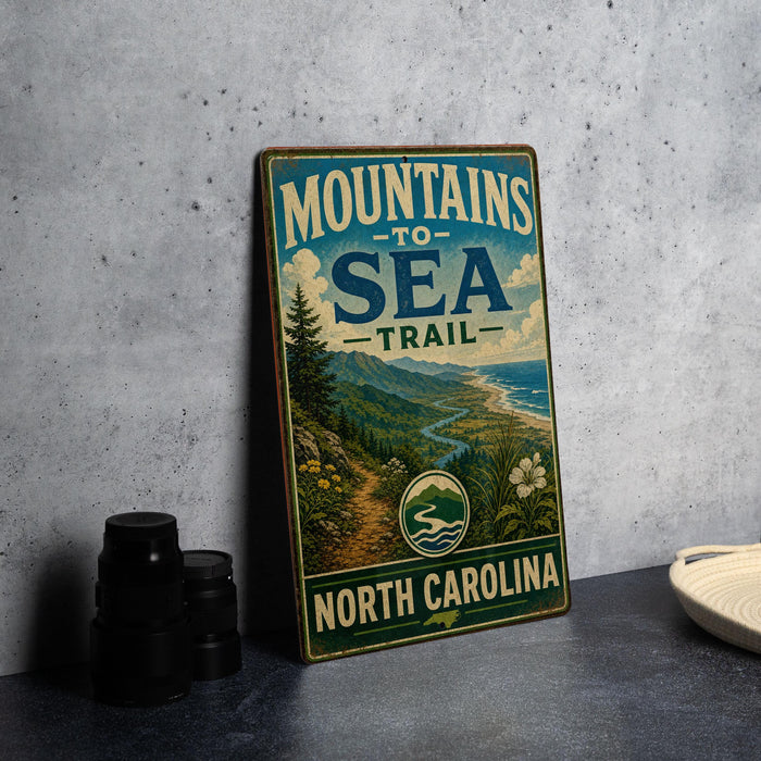 A vintage metal sign with the text "MOUNTAINS TO SEA TRAIL NORTH CAROLINA" and an illustration of a scenic mountain landscape with a river and beach.