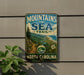 A vintage-style metal sign that reads "MOUNTAINS TO SEA TRAIL NORTH CAROLINA" is displayed on a wall, with a plant to the left of the sign.
