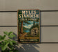 A vintage-style metal sign for the Myles Standish State Forest in Massachusetts, featuring a scenic landscape with pine trees, a lake, and a man in a hat.