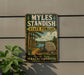 A vintage metal sign for Myles Standish State Forest in Plymouth, Massachusetts, featuring a scenic landscape with pine trees, a lake, and a large rock.