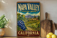 A vintage-style metal sign for Napa Valley, California, featuring a vineyard landscape with grapes and mountains in the background.