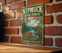 A vintage-style metal sign for the Nipmuck State Forest in Connecticut, featuring a scenic landscape with a lake, pine trees, and a sign indicating hiking and fishing camping facilities.
