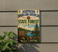 A vintage-style metal sign for the Northern Highland-American State Forest in Wisconsin, featuring a scenic landscape with pine trees, a river, and a sign indicating the location.