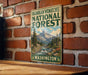 A vintage-style metal sign for the Okanogan-Wenatchee National Forest in Washington, featuring a mountain landscape with a river and pine trees.