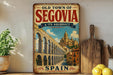 a vintage-style sign for the town of Segovia, Spain, with a picture of the famous Segovia Bridge. The sign also includes the text "Old Town of Segovia & Its Aqueduct" and "SPAIN", indicating the location and the country.