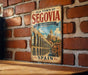 A vintage metal sign with the text "Old Town of Segovia & its Aqueduct" and an illustration of a historical building, likely the famous Segovia Bridge, is displayed on a brick wall.