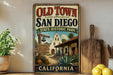 a vintage-style sign for Old Town San Diego State Historic Park, with a sign welcoming visitors to the park. The sign is displayed on a wooden shelf, surrounded by various kitchen utensils and a potted plant.