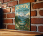 A vintage-style metal sign for the Olympic National Forest in Washington, featuring a scenic mountain landscape with pine trees, a lake, and flowers.