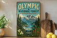 a vintage-style sign for the Olympic National Forest in Washington, with a mountain landscape and a river in the background. The sign is displayed on a shelf next to a wooden cutting board and some potted plants.