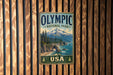 A vintage-style metal sign for Olympic National Park in the USA, featuring a mountain landscape with pine trees and a body of water.