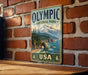 A vintage-style metal sign for Olympic National Park in the United States, featuring a scenic mountain landscape with pine trees and a body of water.