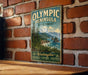A vintage-style metal sign for the Olympic Peninsula State Forest, featuring a scenic landscape with mountains, pine trees, and a body of water.