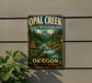 A vintage-style metal sign for Opal Creek Scenic Recreation Area in Oregon, featuring a scenic mountain landscape with a river flowing through it.