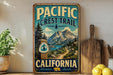 A vintage-style metal sign for the Pacific Crest Trail in California, featuring a scenic mountain landscape and a trail marker.