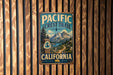 A vintage metal sign for the Pacific Crest Trail in California, featuring a scenic mountain landscape and a path leading to a lake.