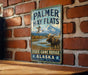 A vintage-style metal sign with the text "Palmer Hay Flats State Game Refuge Alaska Est. 1975" is displayed on a brick wall.