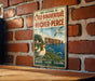 A vintage-style metal sign with a coastal scene, featuring a seagull, cliffs, and water, mounted on a brick wall.