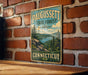 A vintage metal sign for Paugusset State Forest in Connecticut, featuring a scenic landscape with pine trees, mountains, and a lake.